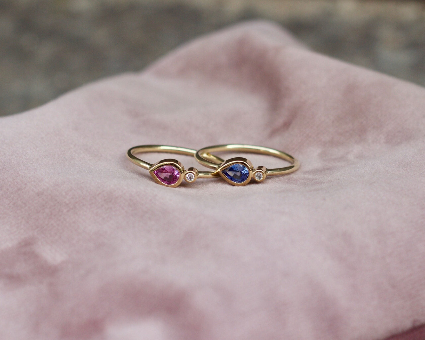 A close-up photo of two stacking gold rings. The top ring features a 0.3Ct blue Sapphire and a brilliant cut 0.02Ct Diamond. The other features a 0.3Ct purple /Pink Sapphire and a brilliant cut 0.02Ct Diamond