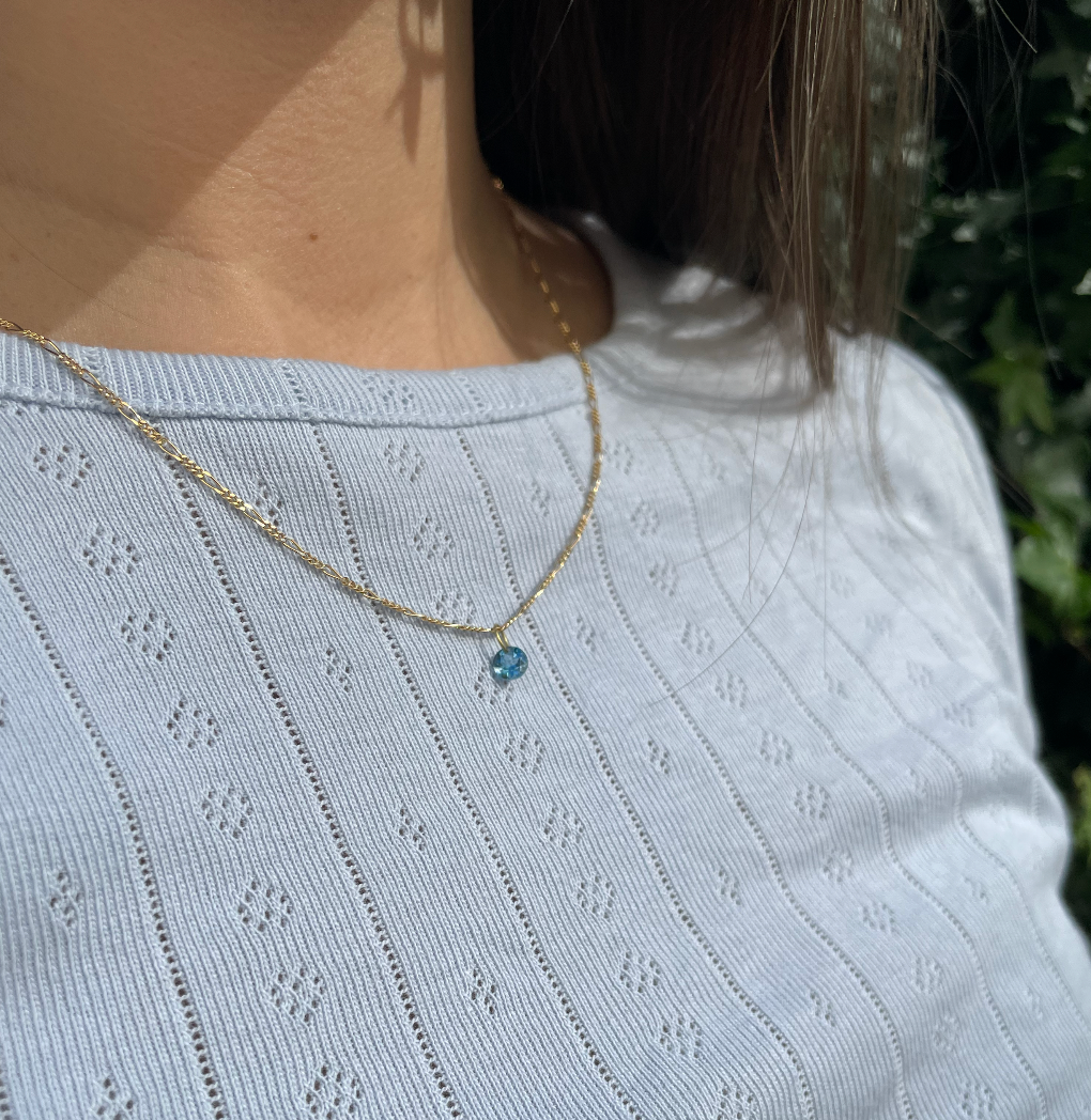18 karat gold Figaro chain. The chain features an alternating pattern of elongated oval links and shorter round links. It is available in a variety of lengths. With a blue tupaz stone pendant.