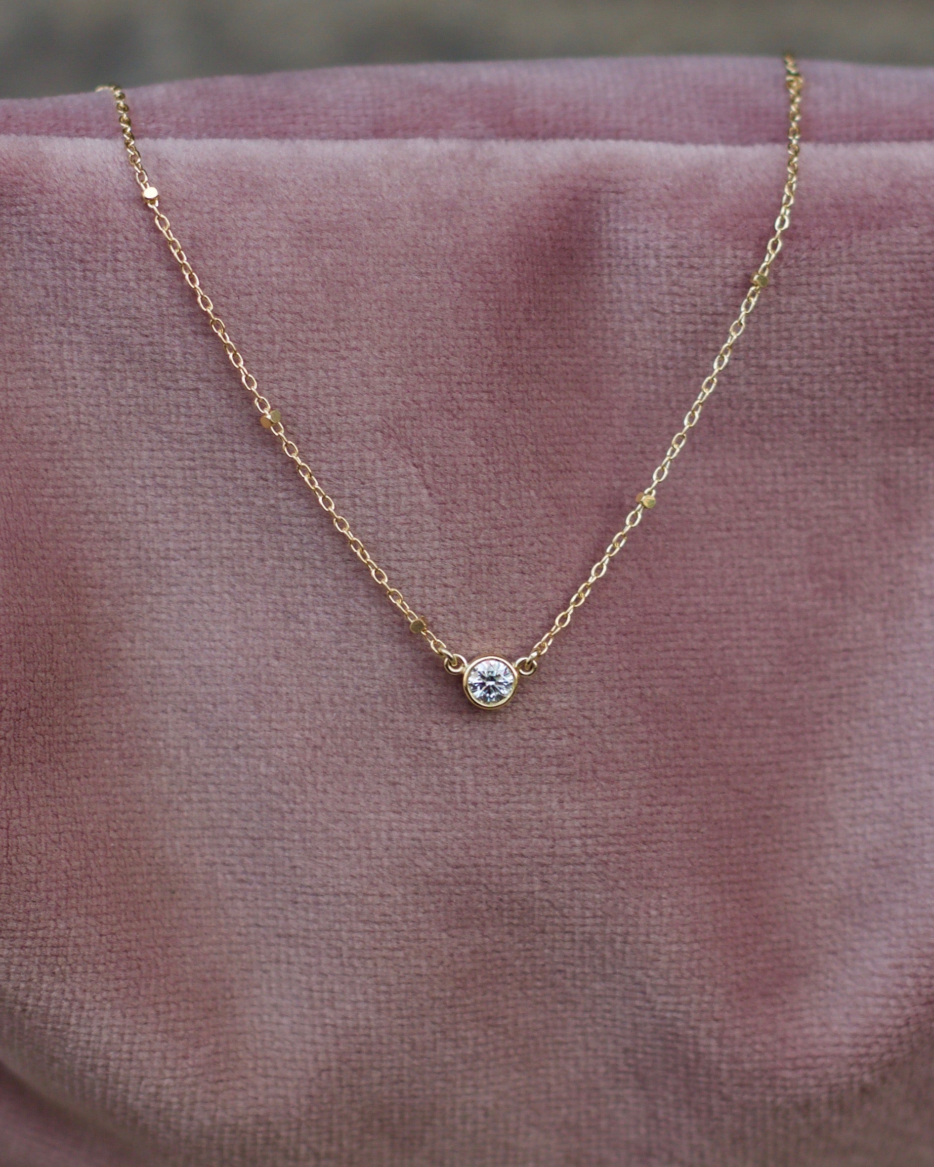 A 18 karat gold necklace featuring a single diamond in a bezel setting. Placed on a pink pillow.